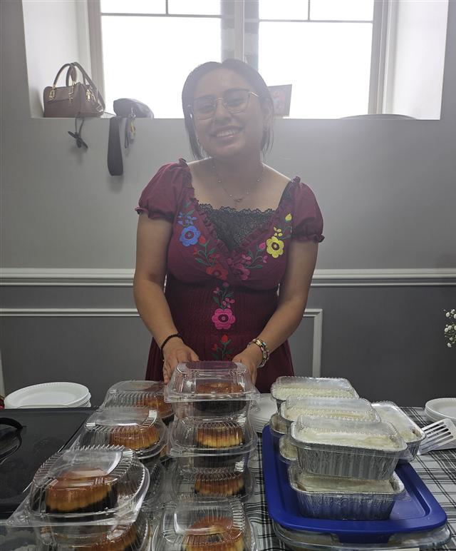 Betsy smiling behind her bake-sale table of homemade flan and tres leches.