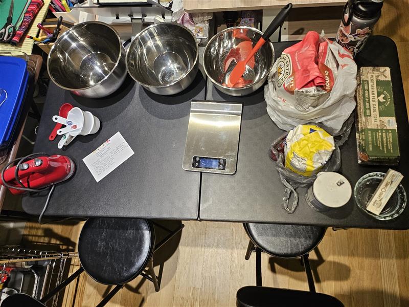 Betsy’s kitchen table set up for baking: empty steel mixing bowls, a digital kitchen scale, measuring cups and bags of flour and sugar.