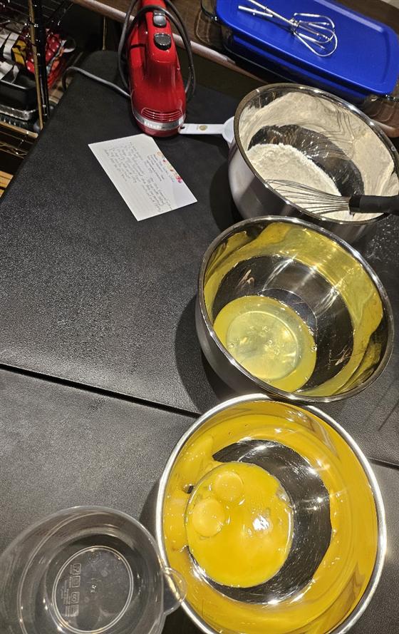 Baking in progress: separated egg whites and yolks in steel bowls, a bowl of flour with a whisk, and a handwritten recipe card.