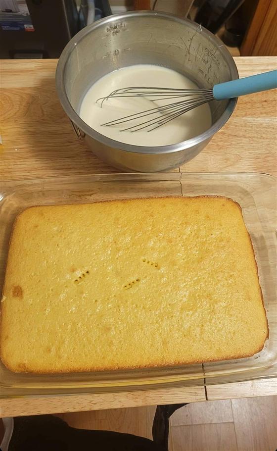 A finished tres leches sponge cake in a glass baking dish, poked with a fork, below a steel bowl of milk mixture with a whisk.